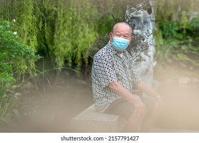 Senior Asian Man Wearing Face Mask, Sitting On A Bench. Outdoor Garden Setting.