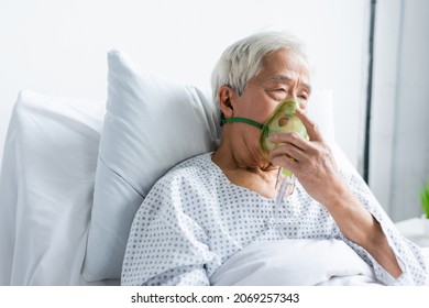 Senior Asian Patient Holding Oxygen Mask While Sitting On Bed In Hospital