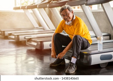 Senior Asian Sport Man Tying Shoelaces On Treadmill In Fitness Gym Ready Walking With Headphone And Water Bottle . Elder Male Exercising , Workout, Training ,healthy ,Retirement ,older, Looking Camera