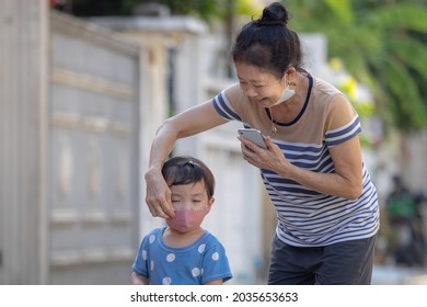 Senior Asian woman (Grandmother) wearing surgical face mask is adjusting her nephew (Grandson) red cloth face mask to prevent coronavirus while walking outside. - Powered by Shutterstock