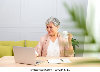 Senior Asian Woman At Phone Works On Her Laptop At Home.