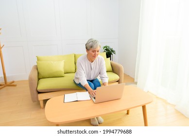 Senior Asian Woman At Phone Works On Her Laptop At Home.