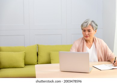 Senior Asian Woman At Phone Works On Her Laptop At Home.