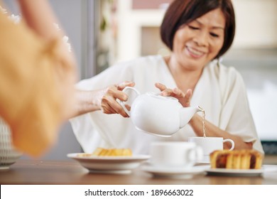 Senior Asian woman sitting at kitchen table and pouring hot tea in cup, selective focus