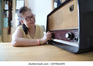 The Senior Asian Woman Trying To Listen Music From The Vintage Radio.