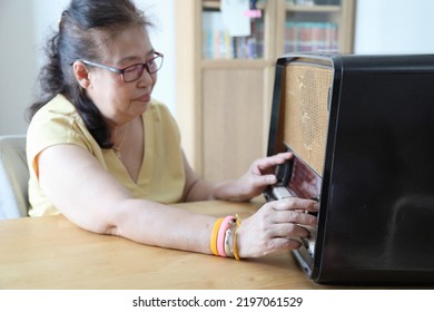 The Senior Asian Woman Trying To Listen Music From The Vintage Radio.