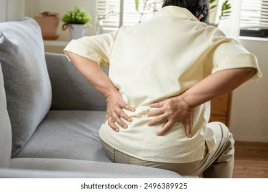 Senior Asian woman using hand pushing on her back because feeling back pain. An elderly person is sitting on a couch, holding their lower back in pain. highlighting discomfort and the need for medical - Powered by Shutterstock