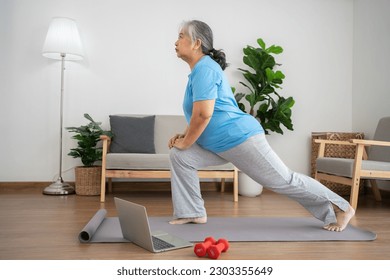 Senior Asian woman watching online courses on a laptop while exercising in the living room at home. Old woman stretching exercises in the living room at home, Concept of workout training online. - Powered by Shutterstock