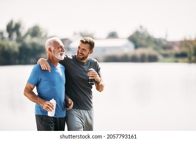 Senior athlete and his adult son talking and drinking water after running by the river in nature. Copy space.
