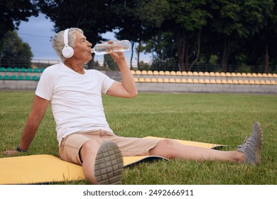 Senior athlete resting listening to music on yoga mat enjoying his outdoor workout at stadium glancing away and drinking fresh cold water during in his outdoor activity - Powered by Shutterstock