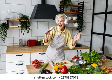 Senior attractive woman listening to music and singing in her headphones in the kitchen. Active lifestyle of mature people, enjoying life, having fun. Using whisk as microphone while cooking food - Powered by Shutterstock