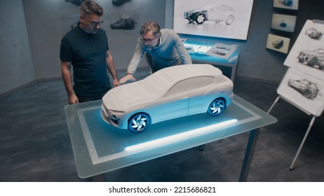 Senior Automotive Developers Make The Design Corrections Looking At The Sculpture Of The Prototype Car Model On A Glass Table. High Tech Office With LED And Car Sketches.