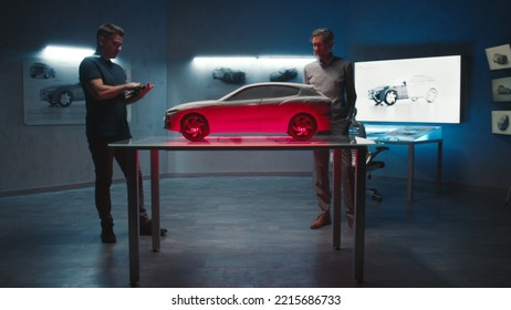 Senior Automotive Engineers Examine The Prototype Car Model Placed On A Glass Table In Different Shades And Colors Of Light. Using A Digital Touchscreen Tablet In A Hightech Car Manufacturing Company.