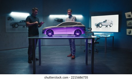 Senior Automotive Engineers Examine The Prototype Car Model Placed On A Glass Table In Different Shades And Colors Of Light. Using A Digital Touchscreen Tablet In A Hightech Car Manufacturing Company.