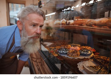 Senior baker examining retail display at his bakery store, looking at delicious donuts and desserts on sale