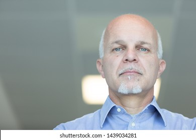 Senior Balding Man With Blue Eyes And A Deadpan Expression Staring Down At The Camera In A Low Angle Head Portrait Indoors