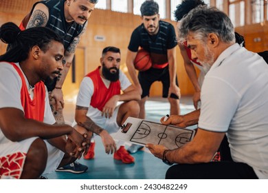 A senior basketball coach is holding clipboard with game tactics while his multicultural team is listening to him and watching game plan on training. Coach is working on game plan with basketball team - Powered by Shutterstock