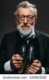 Senior Bearded Citizen In Custom Made Black Suit Holding Bespoke Shoes In Hand, Posing Isolated In Studio.