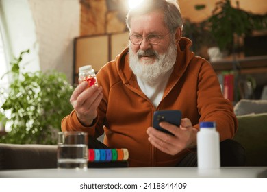 Senior bearded man examining pill bottle while looking at smartphone. Using pill organizer. Online med services, checking prescription. Concept of health and medical care, aging, medicine, treatment - Powered by Shutterstock