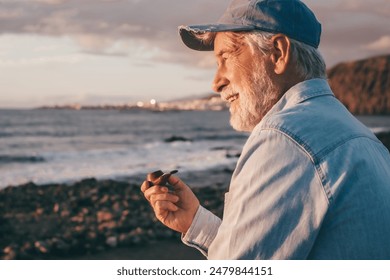 Senior bearded man at the sea beach at sunrise smoking pipe looking at the horizon. Elderly smiling man with cap enjoying vacation or retirement - Powered by Shutterstock