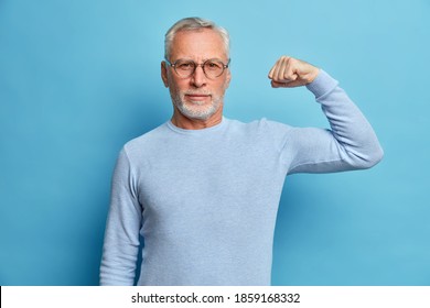Senior bearded man shows muscles after practising bodybuilding wears transparent glasses and basic jumper poses against blue studio background. Look I am very strong and healthy. Perfect biceps