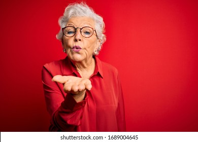 Senior beautiful grey-haired woman wearing casual shirt and glasses over red background looking at the camera blowing a kiss with hand on air being lovely and sexy. Love expression.