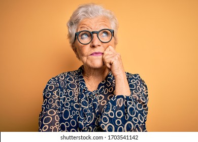 Senior beautiful grey-haired woman wearing casual shirt and glasses over yellow background with hand on chin thinking about question, pensive expression. Smiling with thoughtful face. Doubt concept.