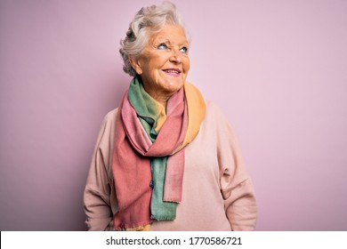 Senior beautiful grey-haired woman wearing casual sweater and scarf over pink background looking away to side with smile on face, natural expression. Laughing confident.