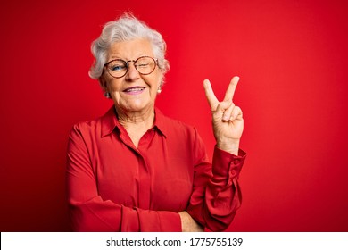 Senior beautiful grey-haired woman wearing casual shirt and glasses over red background smiling with happy face winking at the camera doing victory sign. Number two.