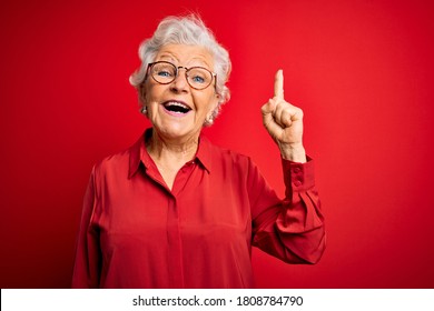 Senior beautiful grey-haired woman wearing casual shirt and glasses over red background pointing finger up with successful idea. Exited and happy. Number one.