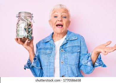 Senior beautiful woman with blue eyes and grey hair holding jar with savings celebrating achievement with happy smile and winner expression with raised hand 