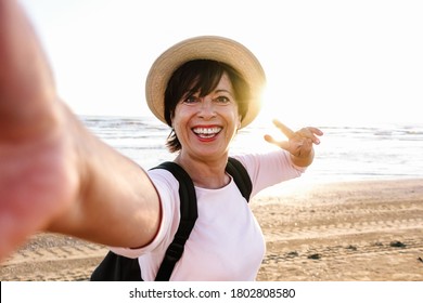 Senior Beautiful Woman With Hat And Backpack Smiling At The Camera Taking A Selfie At The Beach. Older Female Travelling At Holiday.