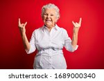 Senior beautiful woman wearing elegant shirt standing over isolated red background shouting with crazy expression doing rock symbol with hands up. Music star. Heavy music concept.