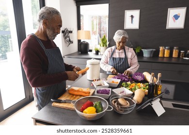 Senior biracial couple in aprons, cutting and peeling vegetables and using tablet in kitchen at home. Senior lifestyle, retirement, communication, food, cooking and domestic life, unaltered. - Powered by Shutterstock