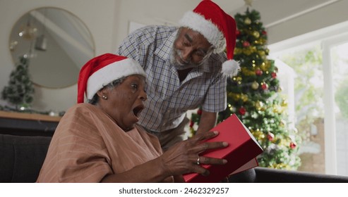 Senior biracial couple celebrating Christmas, grandmother opening gift. Grandfather standing behind with gray hair, both wearing Santa hats - Powered by Shutterstock