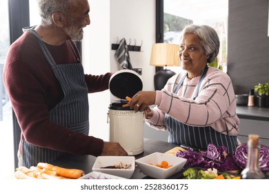 Senior biracial couple wearing aprons composting food waste in kitchen at home. Senior lifestyle, retirement, ecology, food, cooking and domestic life, unaltered. - Powered by Shutterstock