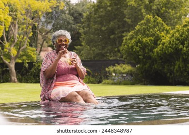 Senior biracial woman enjoys a drink by the poolside, her feet dipped in water, with copy space. She wears a vibrant pink dress and sunglasses, exuding relaxation in a lush garden setting. - Powered by Shutterstock