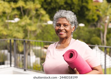 Senior biracial woman with gray hair holds a pink yoga mat outdoors. She smiles warmly, showcasing an active lifestyle and well-being in her golden years. - Powered by Shutterstock