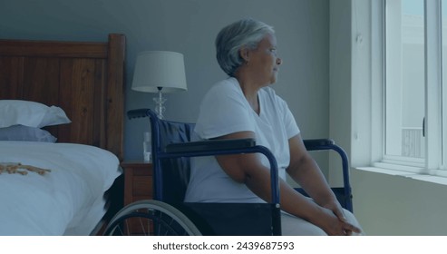 Senior biracial woman sits in a wheelchair at home, gazing outside. She appears contemplative, reflecting on memories or enjoying the view. - Powered by Shutterstock