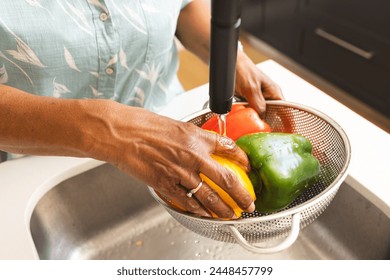 Senior biracial woman washes colorful bell peppers under a kitchen faucet. Her hands show signs of aging, emphasizing the importance of healthy eating at any age. - Powered by Shutterstock