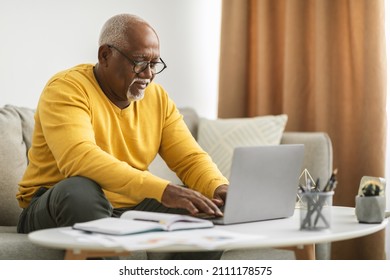 Senior Black Freelancer Man Typing On Laptop Working Online Via Computer Sitting On Couch Indoor. Male Browsing Internet At Home, Wearing Eyeglasses. Freelance Career Concept