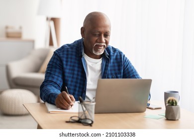 Senior Black Freelancer Man Using Laptop Computer And Taking Notes Working Sitting At Workplace At Home. Older Male Learning Online Writing Something Browsing Internet. Mature People And Gadgets