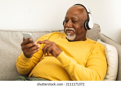 Senior Black Male Listening To Music On Mobile Phone Wearing Wireless Headphones Sitting On Couch Relaxing At Home. Man Listens Audiobook Online Or Using Musical Application Indoor. Great Playlist - Powered by Shutterstock