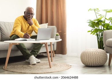 Senior Black Male Using Laptop Working Online And Taking Notes Sitting On Sofa At Home. Mature African American Man Browsing Internet On Computer. Freelance Career And Lifestyle