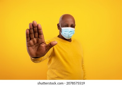 Senior Black Man In Face Mask Gesturing STOP To Camera On Orange Studio Background, Selective Focus. Elderly African American Male Expressing Negativity Towards Coronavirus Pandemic