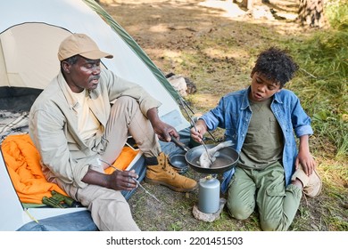 Senior Black Man And His Grandson Cooking Fresh Fish On Frying Pan While Sitting On The Ground By Tent And Enjoying Hike In The Forest