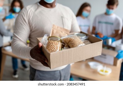 Senior black man holding box with donations food, cans and packages with grains and pasta, cropped. Group of millennial volunteers packing boxes with products for poor and homeless people