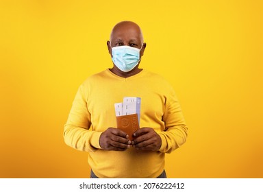 Senior Black Man In Medical Mask Holding Plane Tickets And International Passport On Orange Studio Background. Elderly African American Guy Traveling Overseas During Coronavirus Lockdown