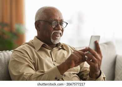 Senior Black Man Texting On Smartphone And Using Mobile Application Wearing Eyeglasses Sitting On Sofa At Home. Male Scrolling Reading News Feed In Social Media And Browsing Internet Via Phone