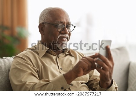 Senior Black Man Texting On Smartphone And Using Mobile Application Wearing Eyeglasses Sitting On Sofa At Home. Male Scrolling Reading News Feed In Social Media And Browsing Internet Via Phone Stock foto © 